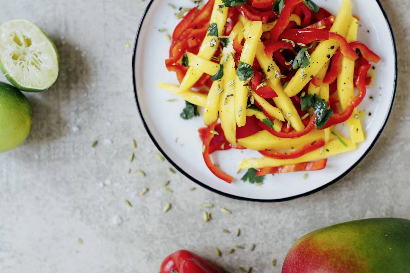 Mango-Fenchel-Salat auf einem weißen Teller mit blauem Rand, daneben eine Mango und halbierte Limetten