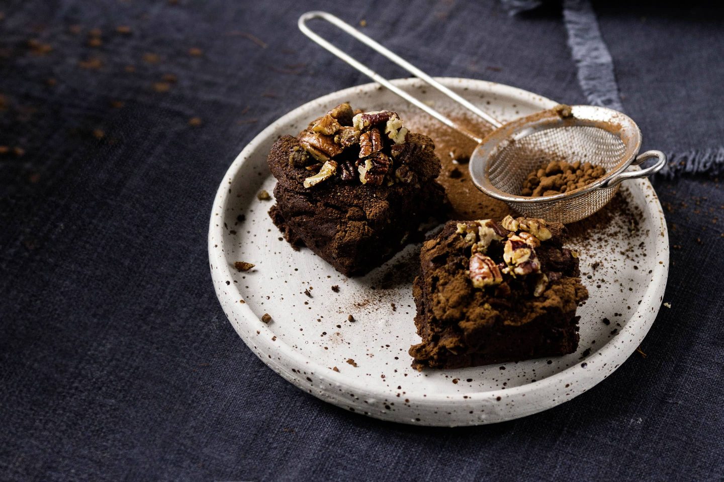 2 Stück Gewürz-Brownies mit Karamell-Pekannüssen auf einem hellen Teller mit Kakaopulver bestreut