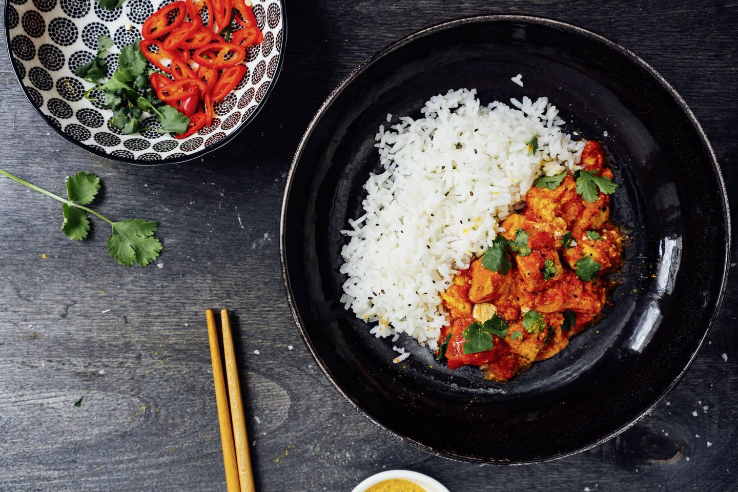 Neapesisches-Cardamom-Huhn mit Reis auf einem tiefen weißen Teller mit Essstäbchen und einer schwarz-weiß-gemusterten Schale mit fein geschnittener frischer Chili