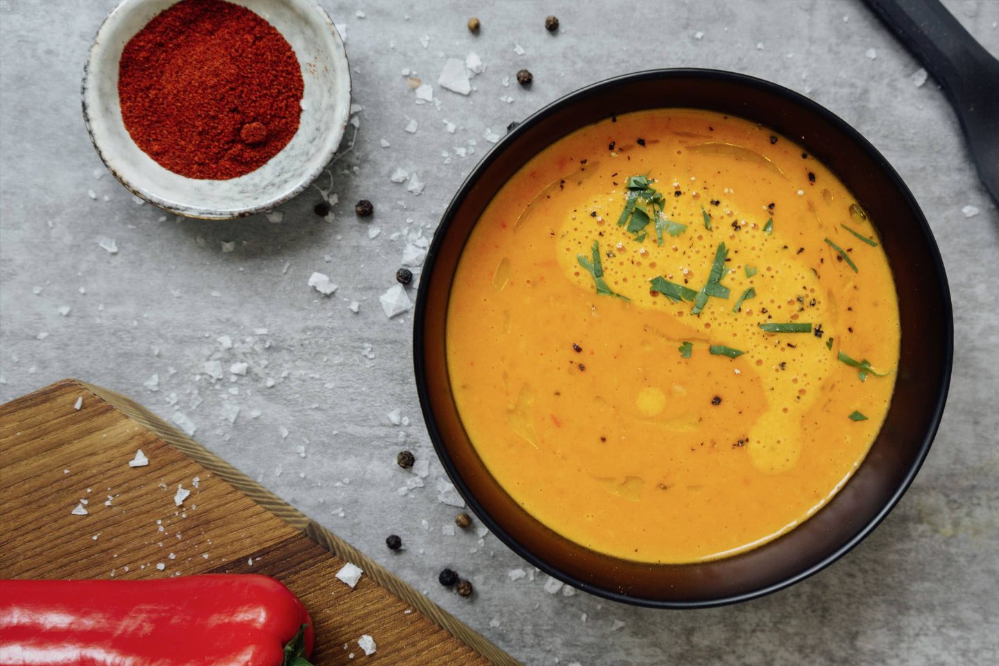 Paprikaschaumsuppe in einer schwarzen Schüssel, daneben ein Brett mit Paprika und ein Schälchen mit Cayenne-Pfeffer