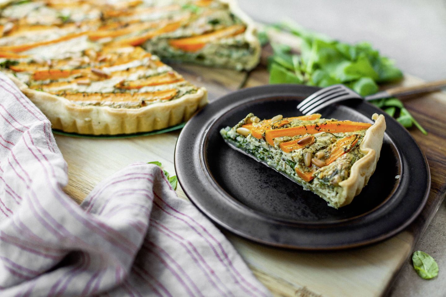Ziegenkäse-Quiche mit Blattspinat und Karotten und ein Stück davon auf einem schwarzen Teller