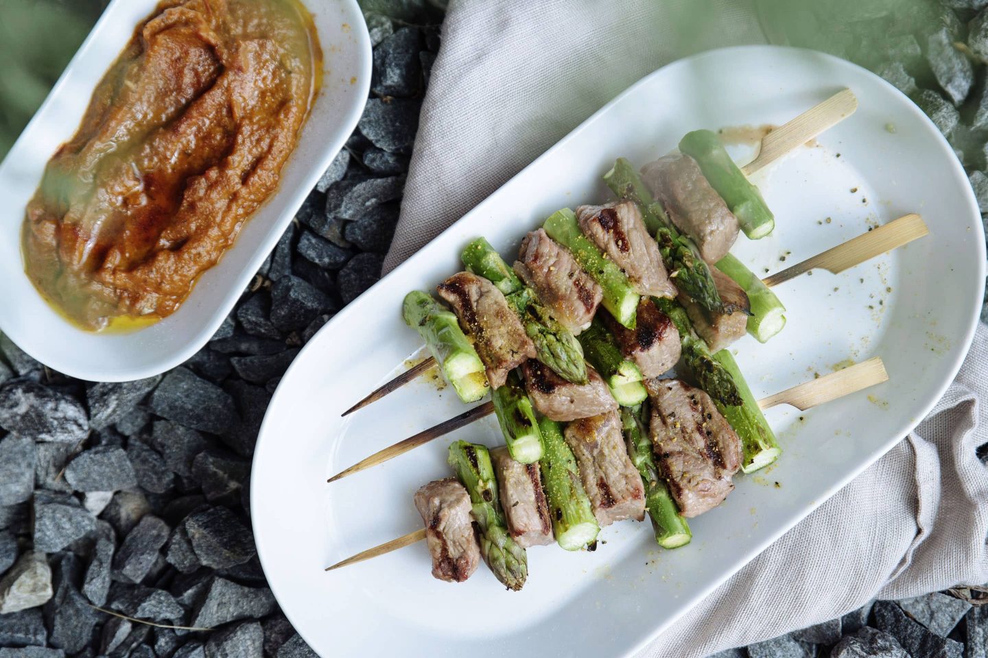 Steak-Gewürzsalz-Spieße mit Spargel auf einem weißen länglichen Teller und Ajvar dazu in einem separaten Schälchen