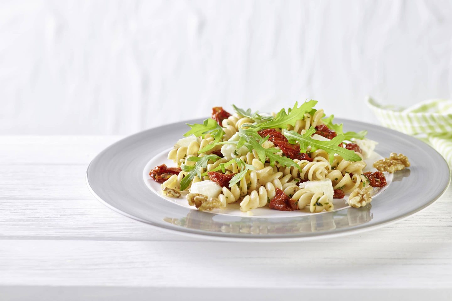 Vollkorn Fussili mit getrockneten Tomaten, Feta und gerebeltem Basilkum auf einem hellen Teller mit frischem Rucola garniert