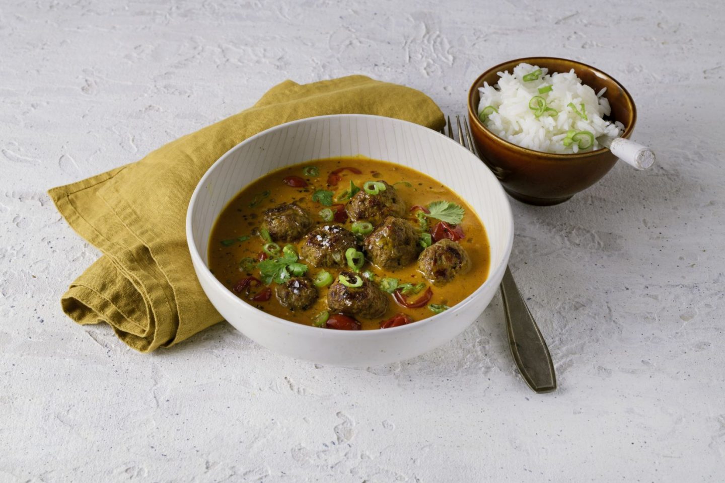 Meatballs in Kokos-Tomatensauce mit Jungzwiebelringen bestreut und in einer Schüssel angerichtet, mit Reis und Serviette