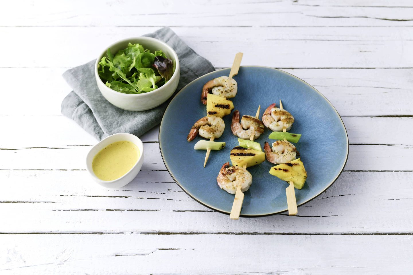 3 Stück Grill Fisch Garnellenspiesse mit Ananas und Curry-Dip auf einem blauen Teller mit einem Schälchen Blattsalat daneben