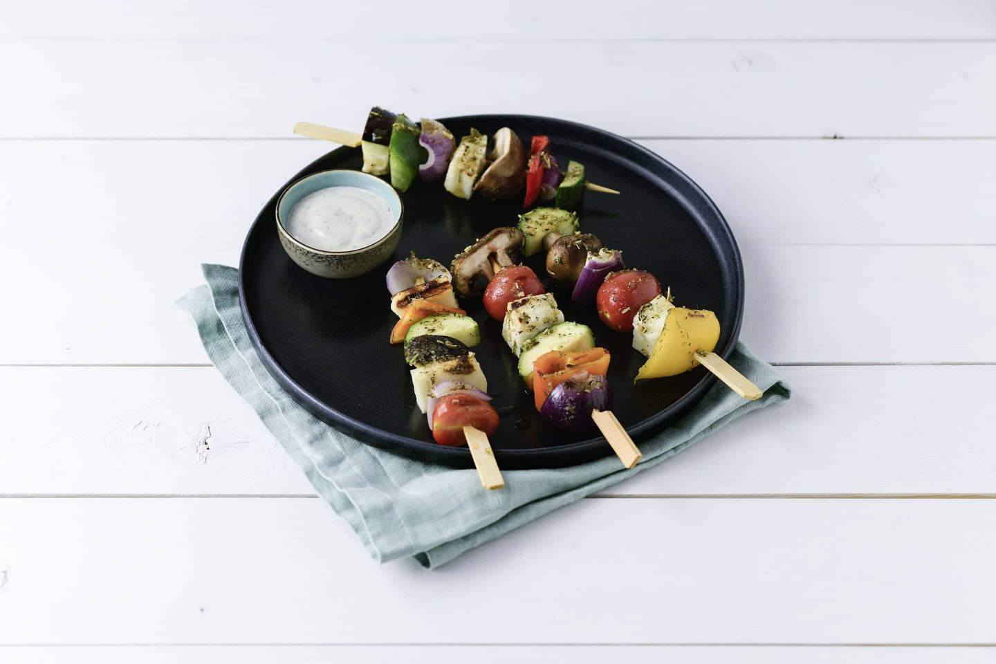 Grill-Gemuese Gemuesespiesse mit Knoblauch-Dip auf einem schwarzen Teller