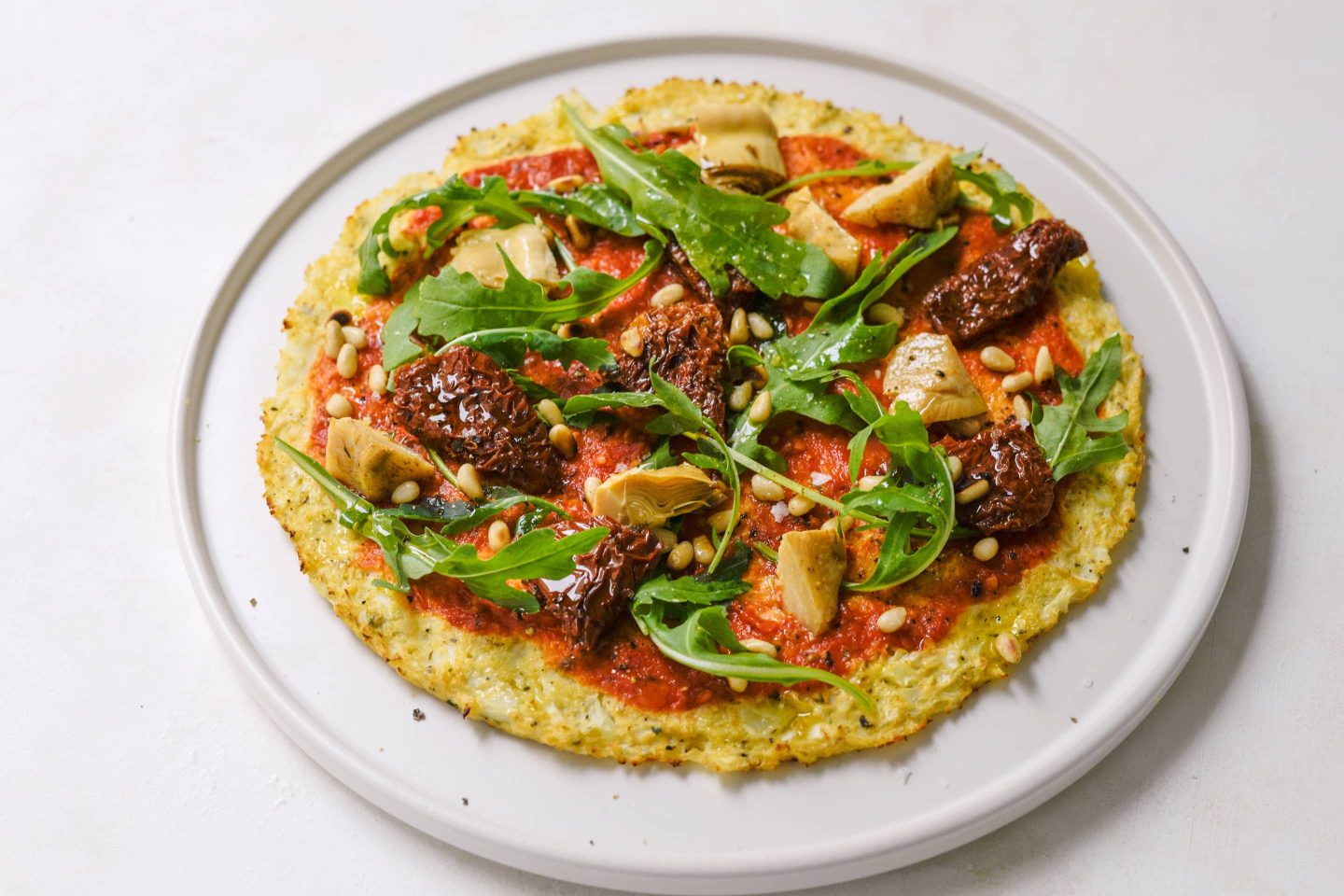 Karfiolpizza Karfiolpizza mit Rucola, getrockneten Tomaten und Artischocken auf einem weißen Teller