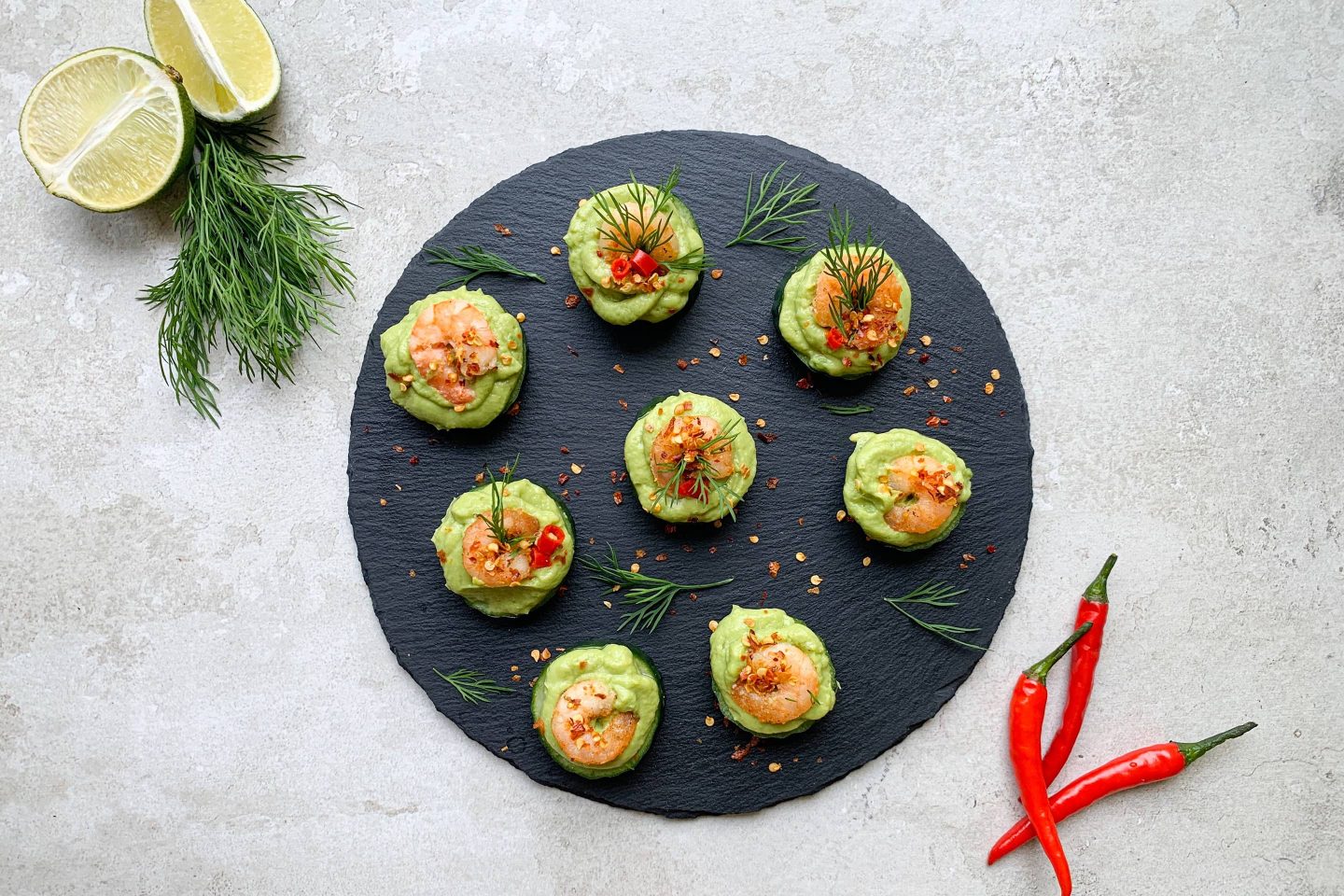 Garnelen-Avocado-Türmchen mit frischem Dill garniert auf einem runden Schieferteller angerichtet, daneben halbierte Limetten und frische Chilischoten