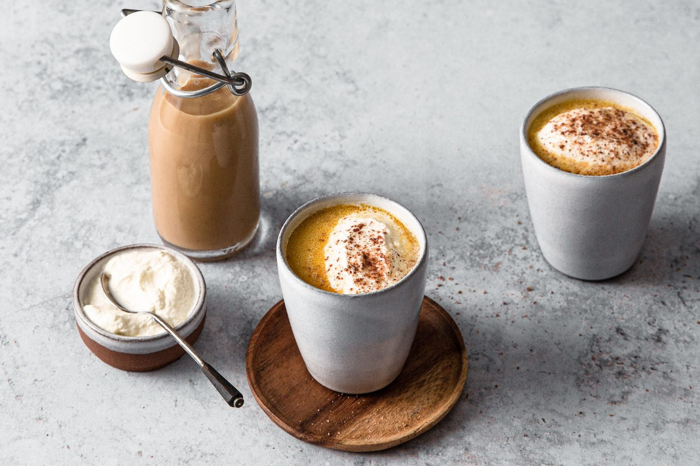 2 Becher mit Chai Coffee Eierlikoer die auf Holztellerchen stehen, eine kleine Schale mit Schlagobers und Löffelchen und eine Flasche mit dem Eierlikör im Hintergrund. Alles steht auf einer grauen Platte.