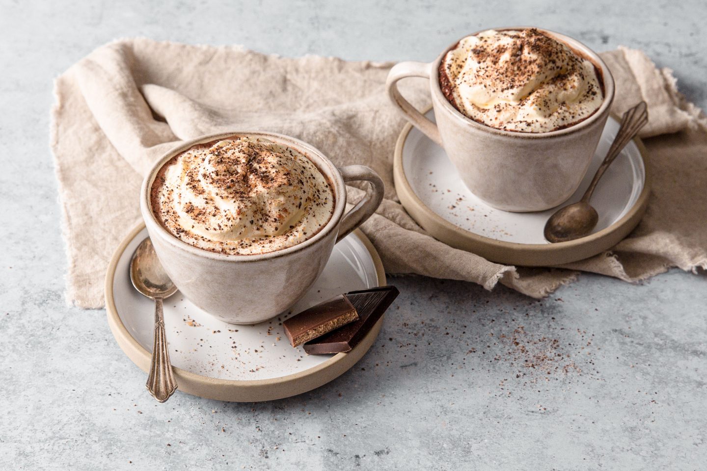 2 Tassen mit heißer Schokolade und Schlagoberhäubchen mit Cacao Kiss Topping aus der Mühle, mit Schokostückchen auf der Untertasse angerichtet