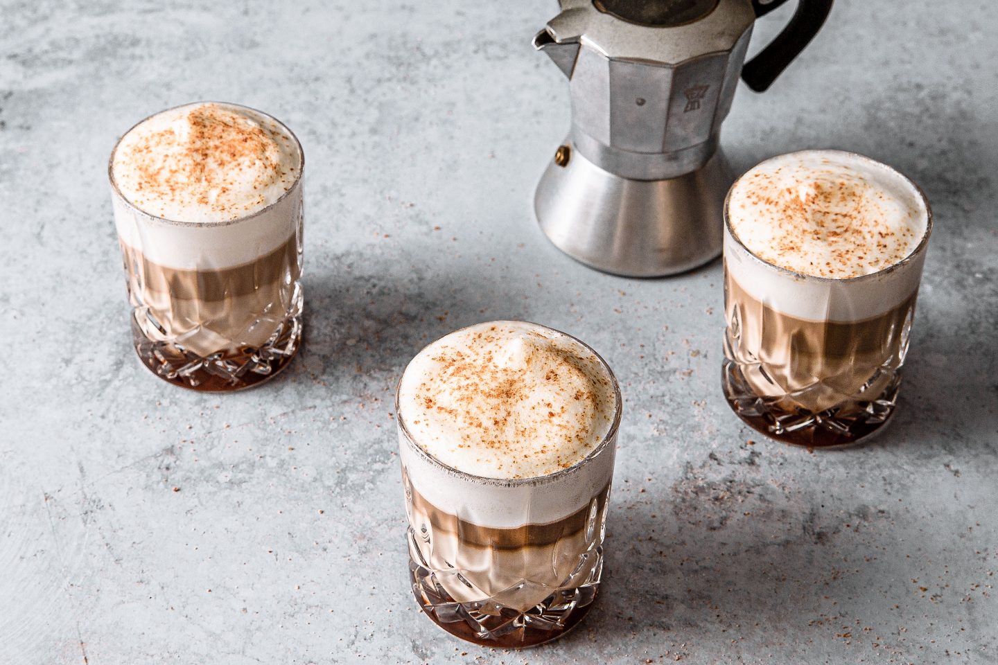 3 Gläser auf einer grauen Oberfläche, die mit Salted Caramel Macchiato gefüllt sind und mit Salted Caramel aus der Mühle getoppt. Daneben platziert ist eine kleine Alu-Espressomaschine für den Herd