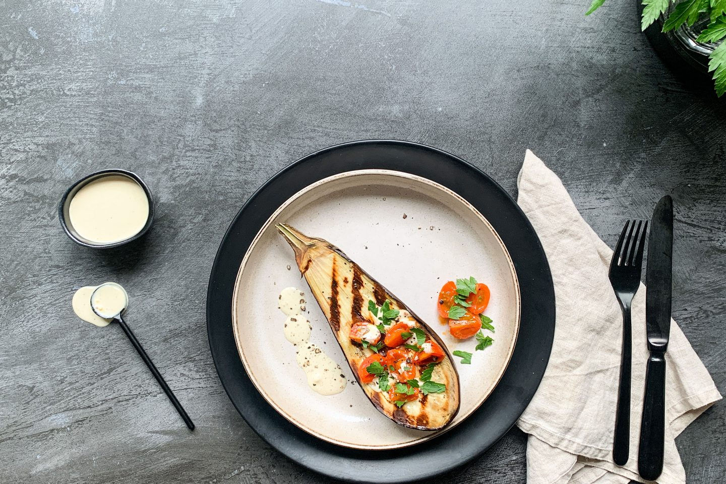 Foto: Gegrillte Melanzanihälfte mit geschmolzenen Tomaten auf einem hellen Teller angerichtet und mit etwas Erdnusssauce beträufelt