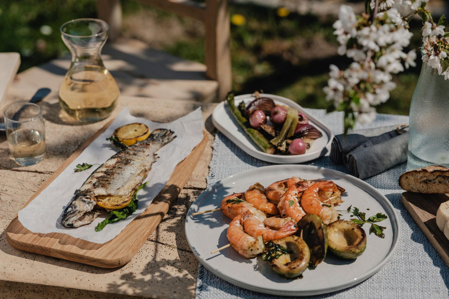 verschiedene Grillgerichte von Forelle bis Grillgemüse und Shrimp-Spieß auf einem Holztisch im Garten