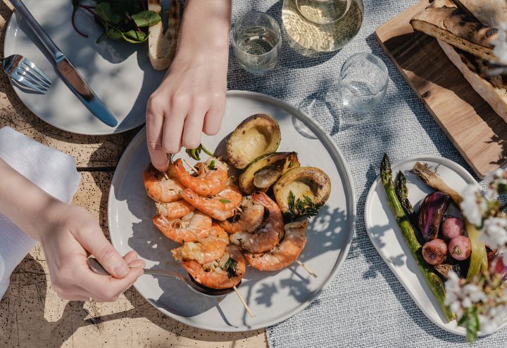 Hände nehmen sich gerade einen gegrillten Garnelenspieß vom Teller