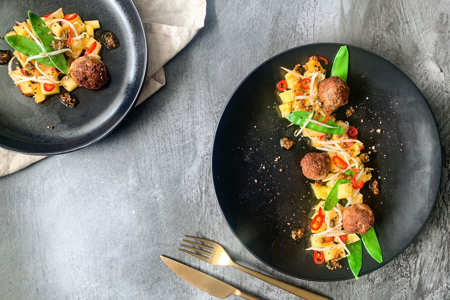 Foto: Salat mit frischen Erbsenschoten, frittierten Kokos-Huhn-Bällchen und Ananas auf einem schwarzen Teller mit goldenem Besteck angerichtet