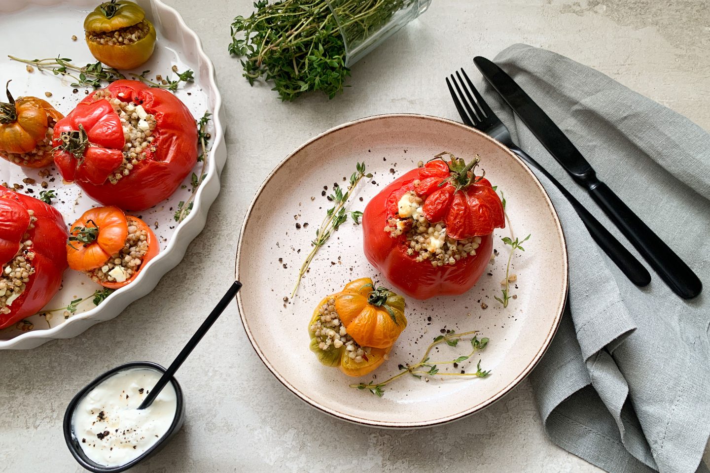 2 gebackenene Ochsenherztomaten mit Buchweizenfülle auf einem beigen Teller mit weiteren Tomaten in einer Auflaufform, frischem Thymian und Sauce