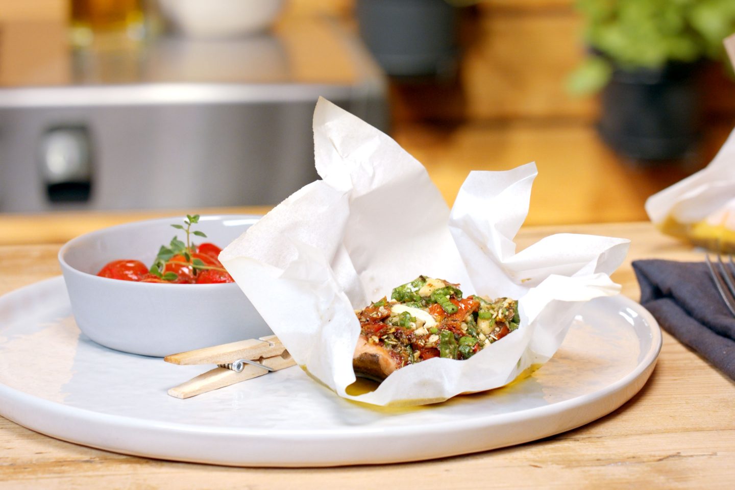 Lachsforelle in einem Tascherl aus Backpapier mit Grill Fisch Grillgewürz, Erdnüssen und Jungzwiebel getoppt, neben einem Schälchen mit Schmelztomaten