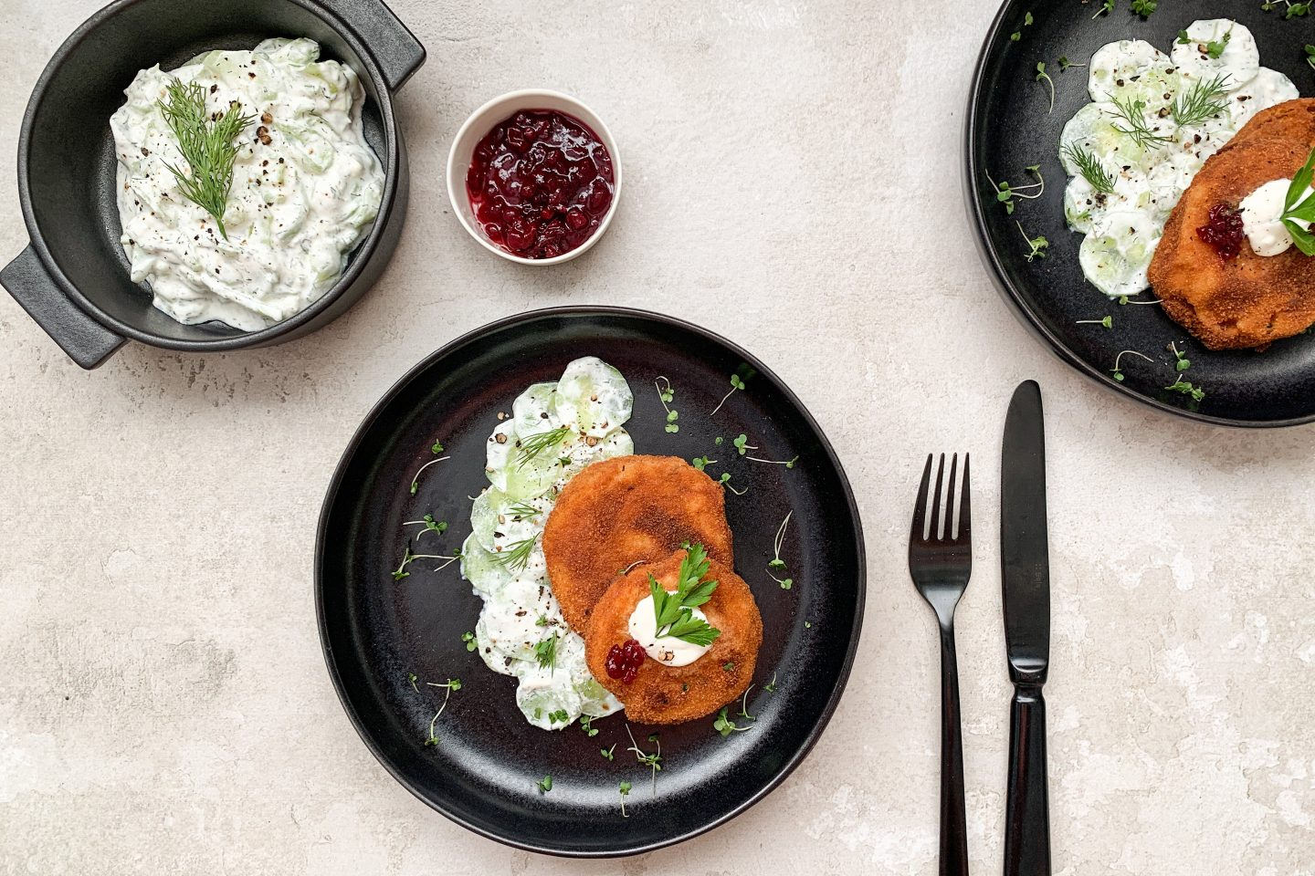 Kohlrabi Schnitzel mit cremigem Gurkensalat auf einem schwarzen Teller mit Preiselbeermarmelade