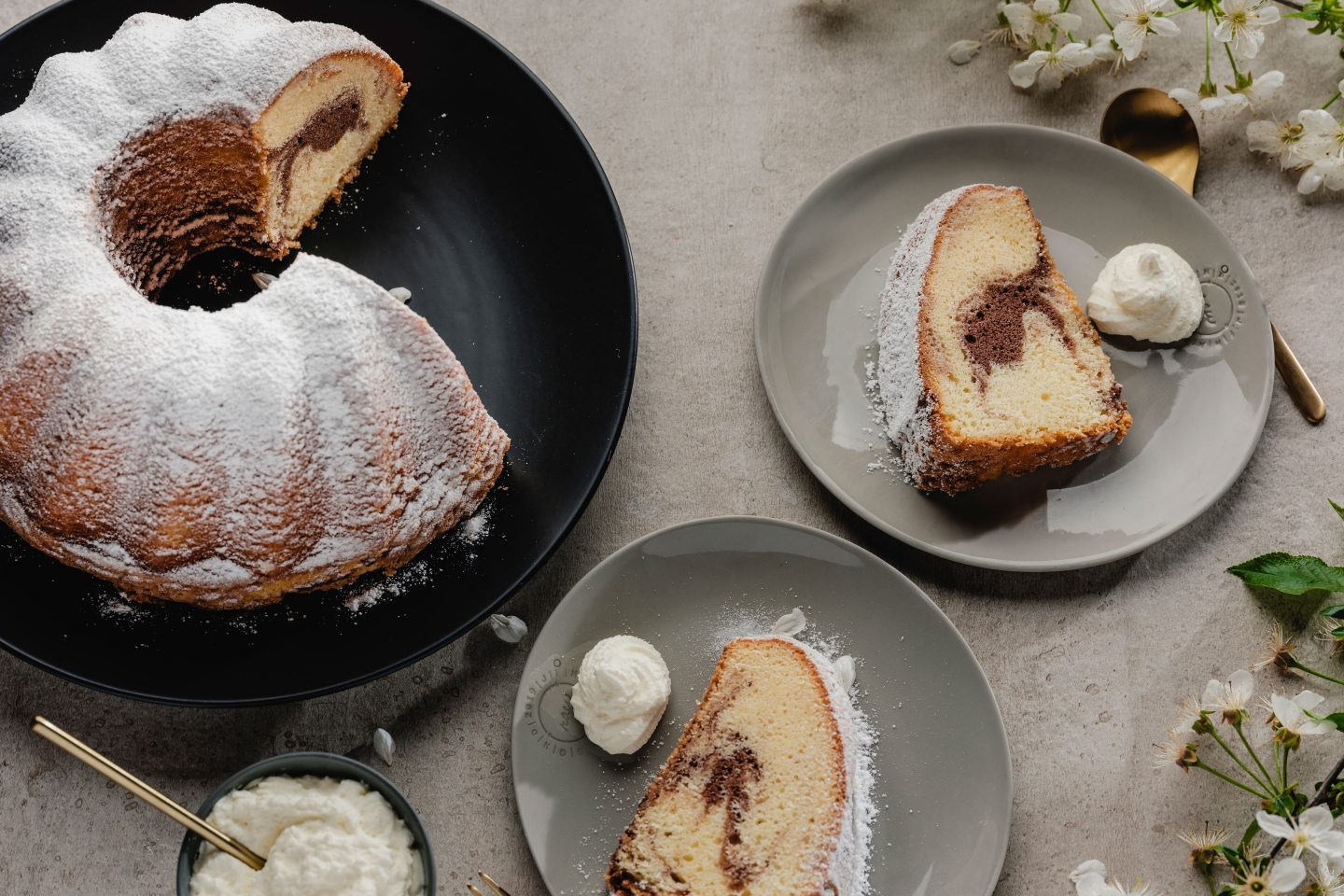 Marmor-Guglhupf auf einem schwarzen Teller und 2 Stück Kuchen auf kleinen Tellern mit Schlagobers, goldenen Gabeln und Apfelblüten
