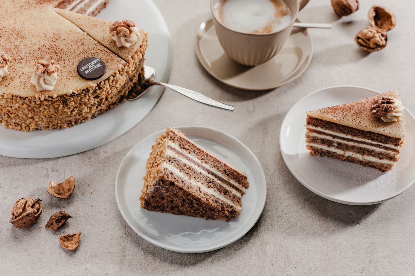 2 Stück Nusstorte mit Haselnusskrokant auf kleinen Tellern mit einer Tasse Cappuccino