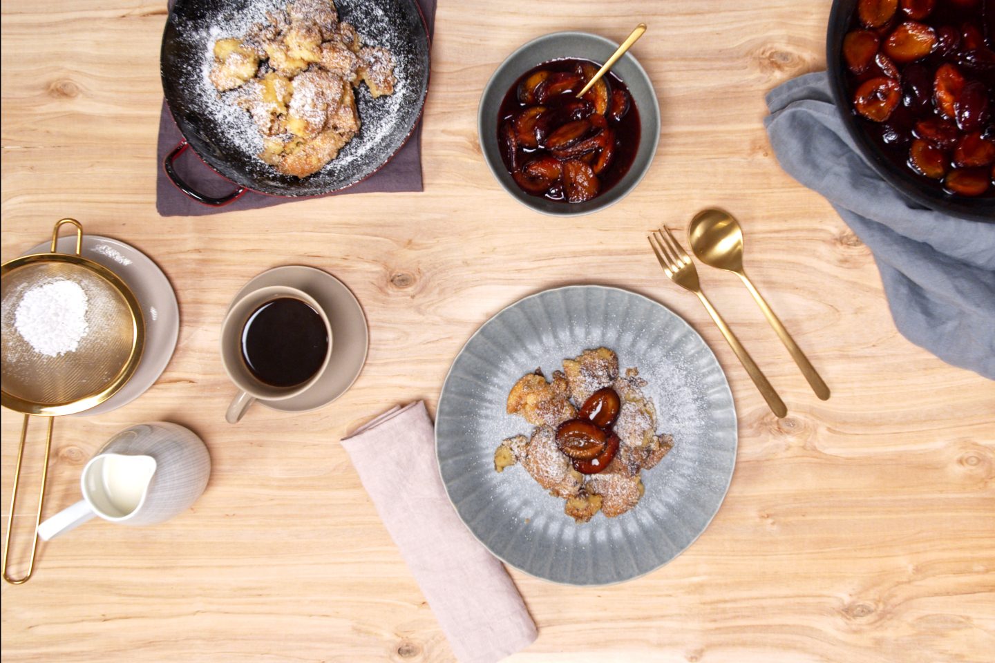 Kaiserschmarrn auf einem blauen Teller mit Staubzucker bestreut, dazu gibt es Pfefferzwetschken in einem blauen Schüsselchen und einer Pfanne