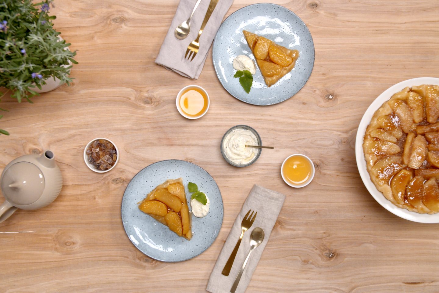 2 Stück Tarte Tatin auf hellblauen Tellern, daneben der ganze Kuchen auf einer weißen Etagere
