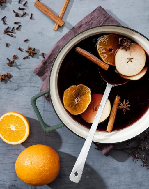 Gluehwein in einem Topf mit Henkeln und Schöplöffel, daneben liegen Gewürze und Orangen auf der Arbeitsfläche
