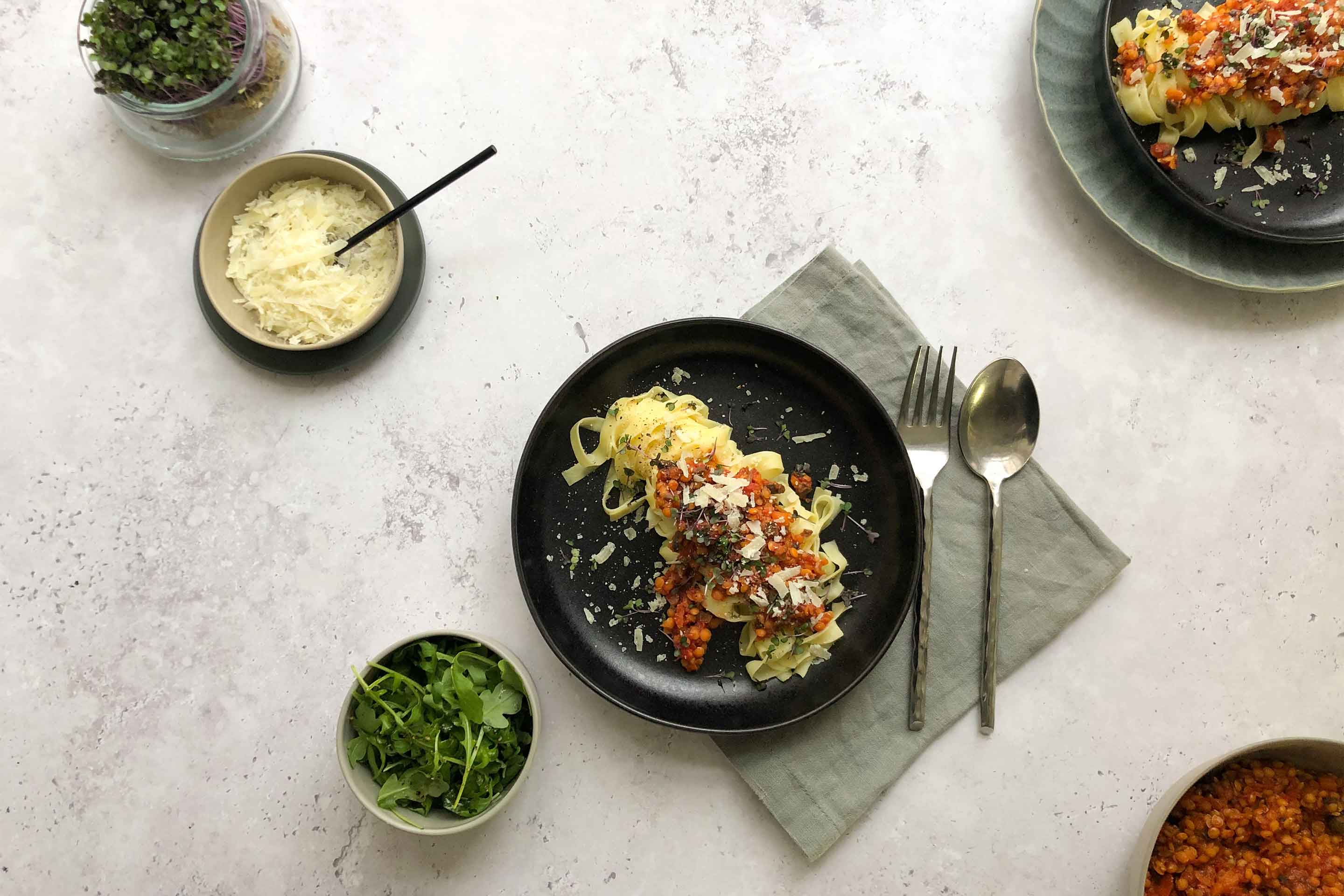 Linsen Bolognese auf Linguine; auf einem schwarzen Teller platziert daneben eine Schüssel mit geriebenem Parmesan