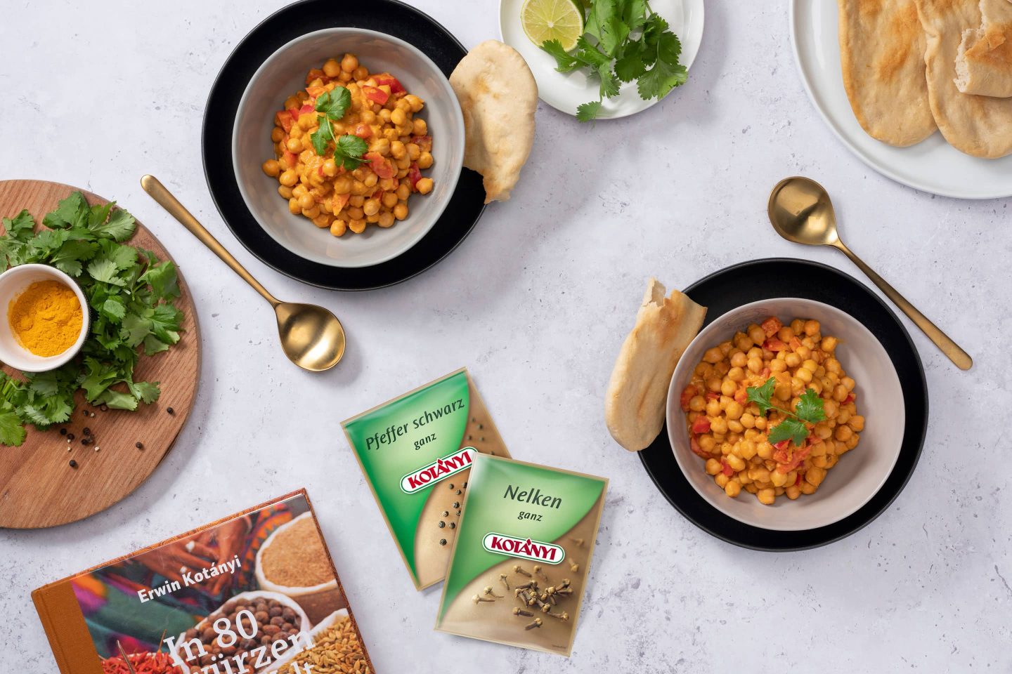 Kichererbsen-Curry in zwei cremefarbenen Schüsseln mit Koriander garniert, daneben ein Teller mit Naan und zwei Kotányi-Biefverpackungen aus den 2000er Jahren.