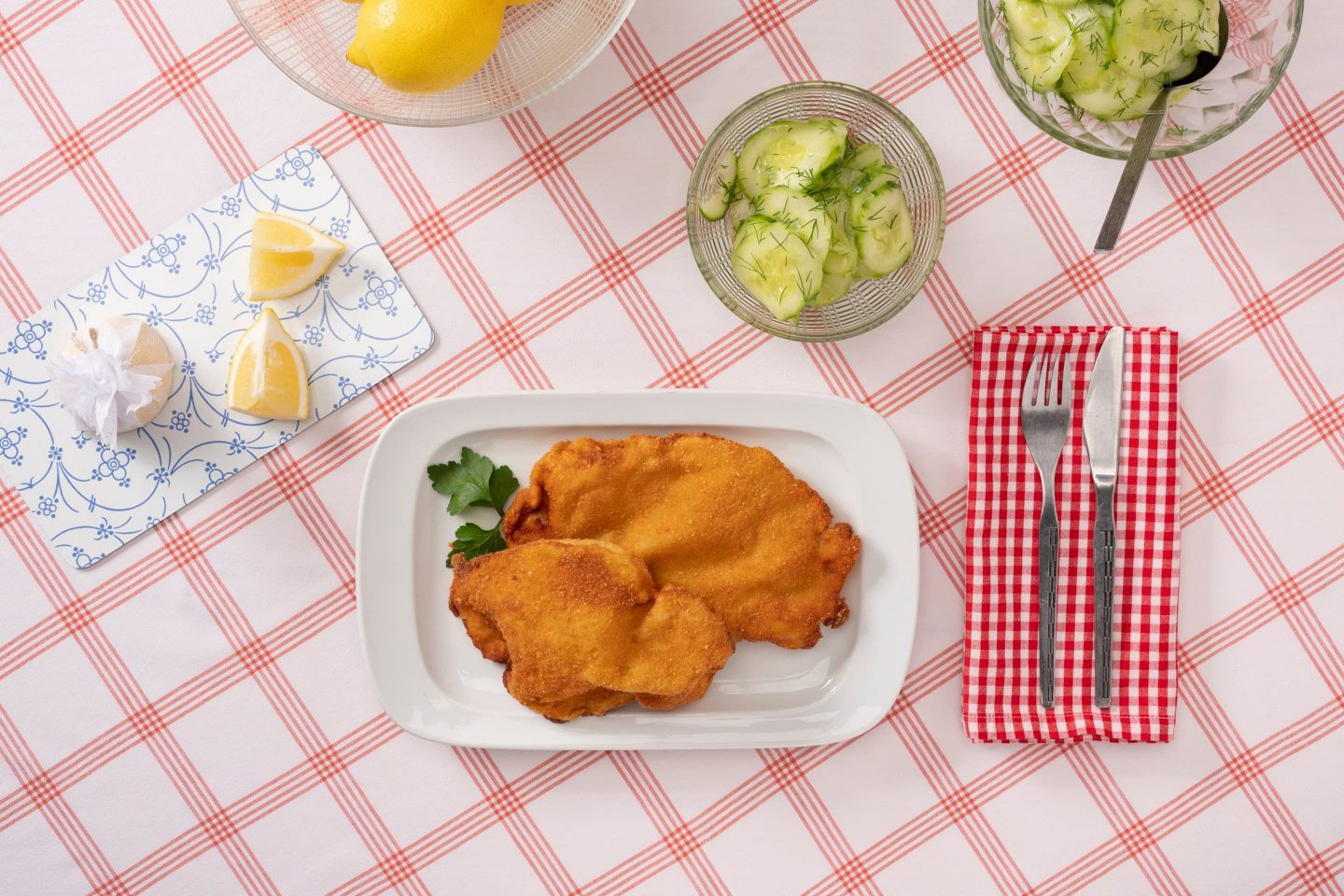 2 Wiener Schnitzel auf einem rechteckigen Teller, mit Gurkensalat in einem gläsernen Schälchen, auf rot-weiß-karierter Tischdecke