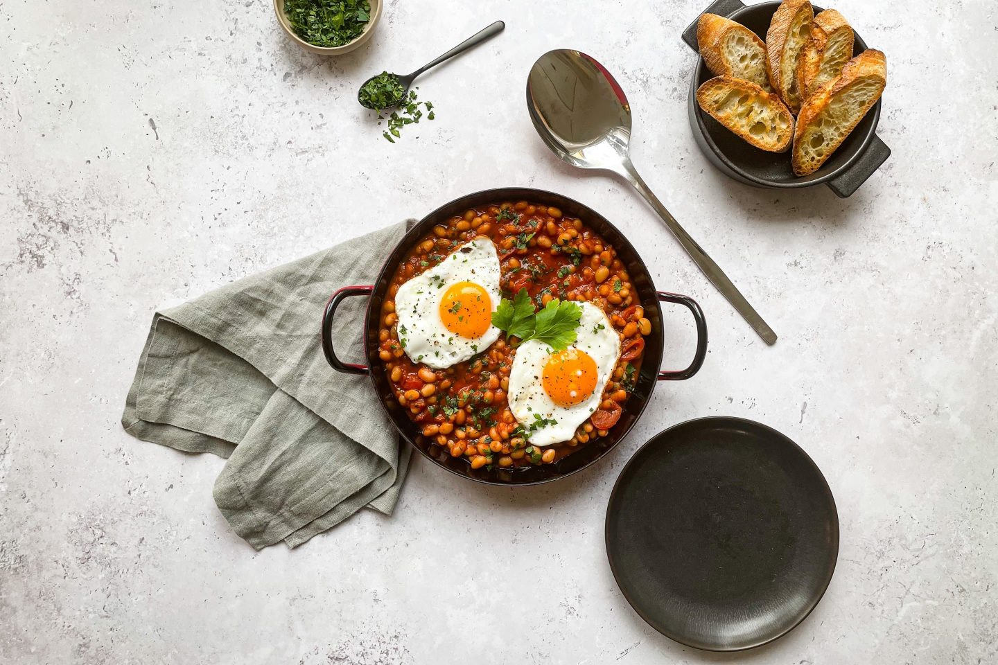 Eine heiße Pfanne Baked Beans und Spiegelei mit Brot, Teller und Serviette.