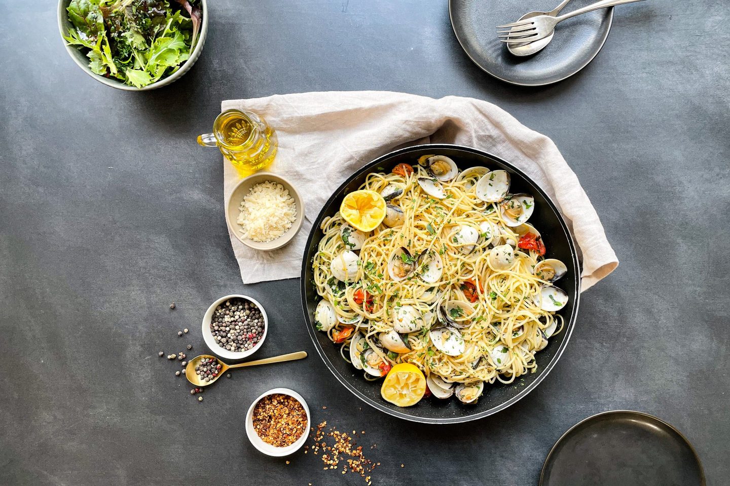 Eine Schüssel mit Pasta Vongole auf einem Geschirrtuch mit Salat und Gewürzen.