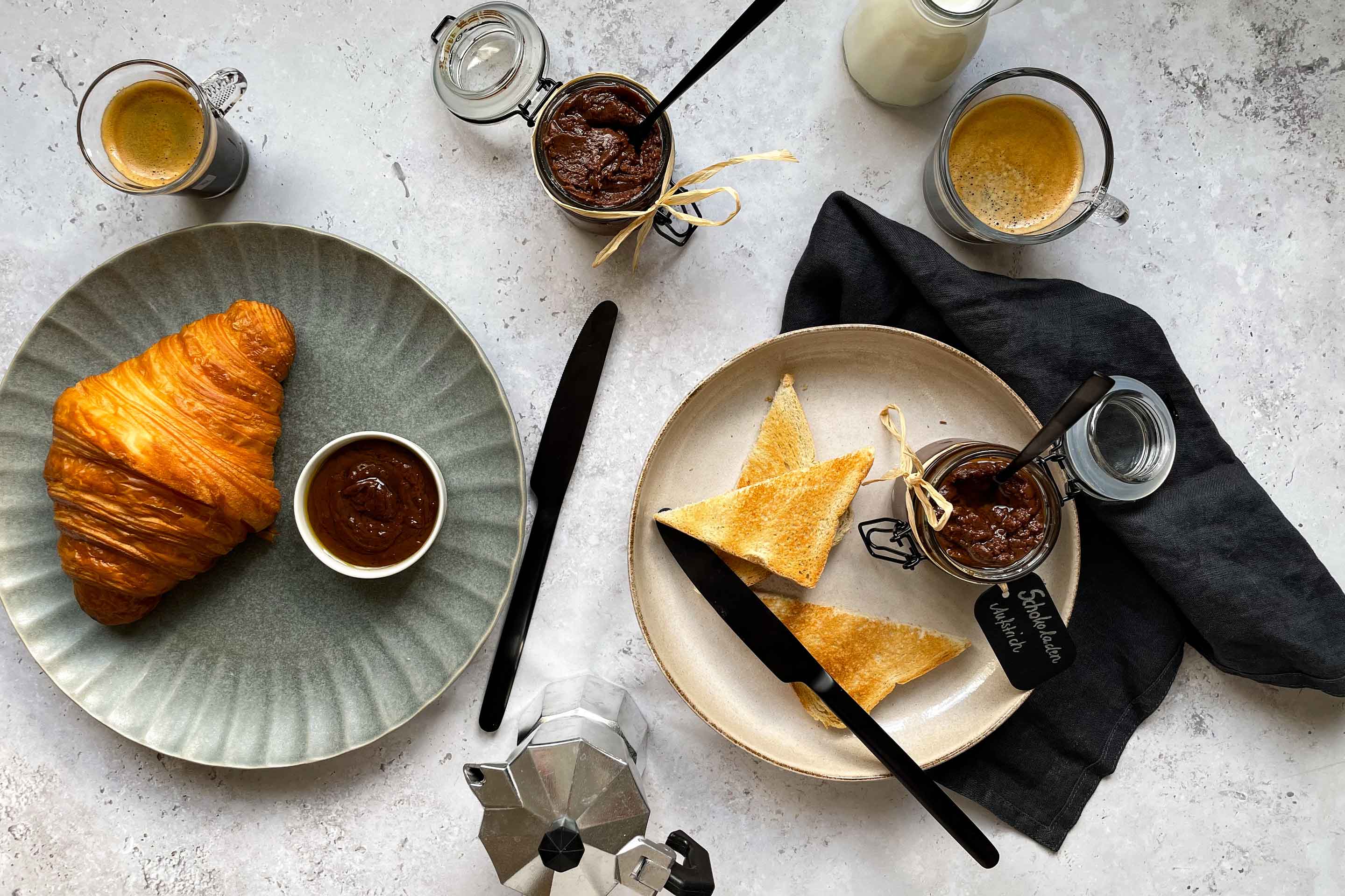 Ein Frühstückstisch mit Kaffee, Croissant, Toasts und Gläschen gefüllt mit Schokoaufstrich.