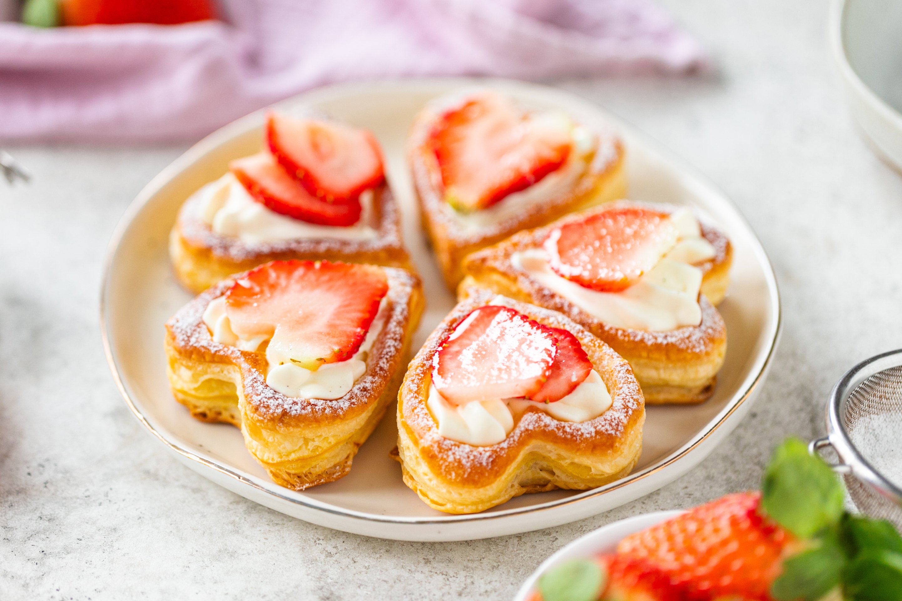 Heart-shaped puff pastries topped with cream and sliced strawberries, dusted with powdered sugar.